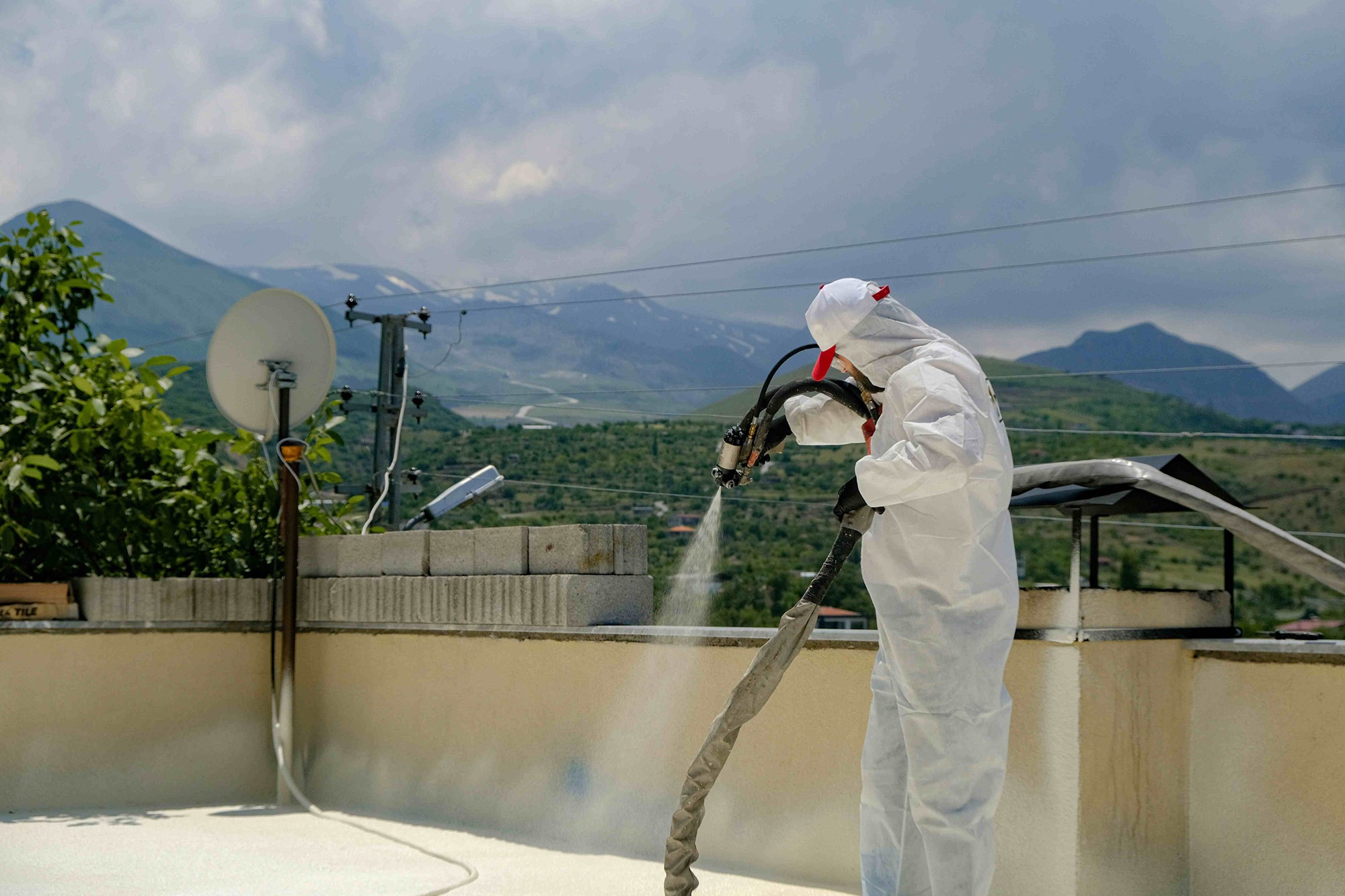 A person in protective gear sprays a surface with a hose on a rooftop, with mountains in the background. This demonstrates a construction or maintenance process.