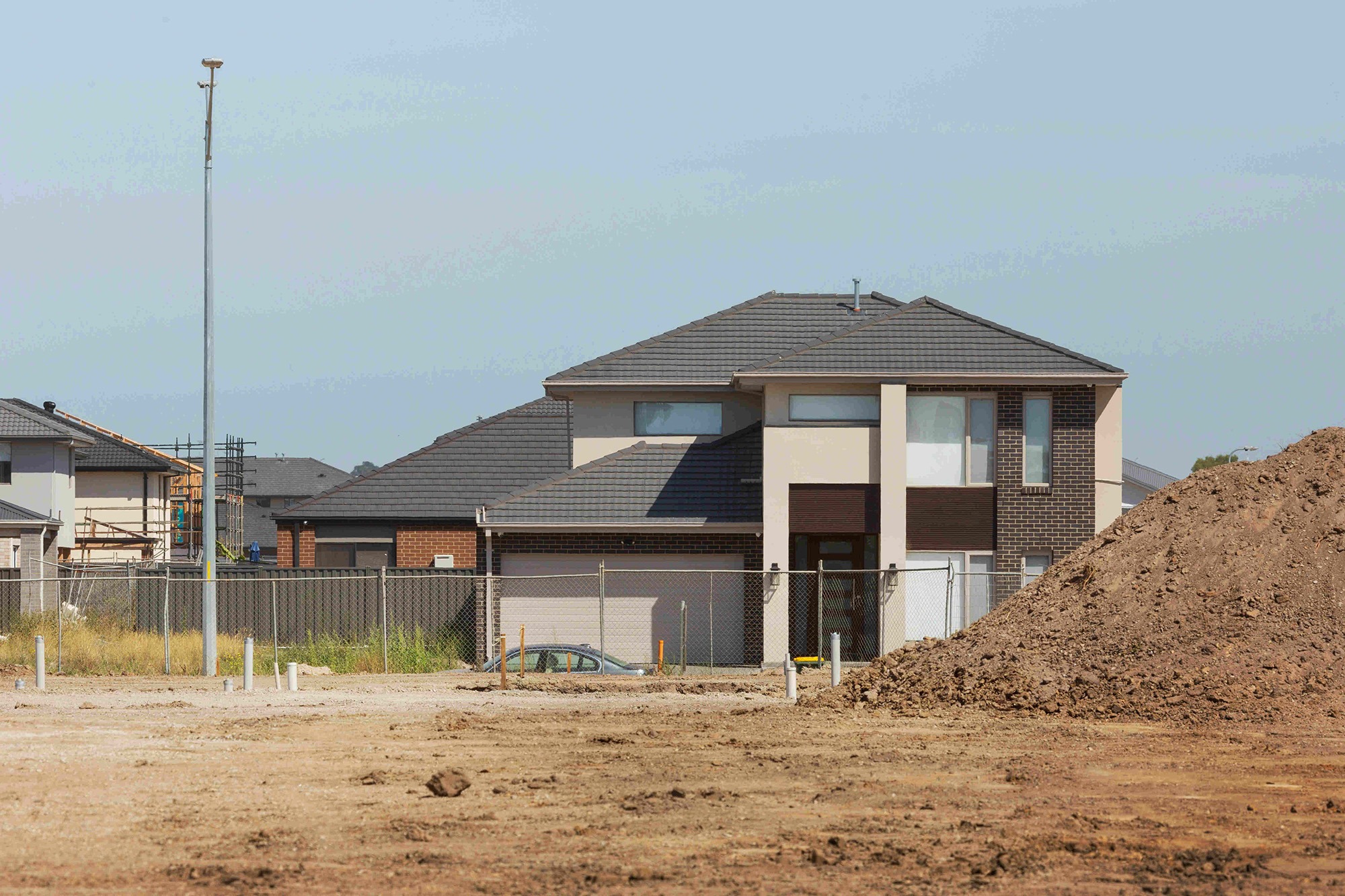 Newly constructed house stands near an empty lot with dirt mounds, highlighting ongoing development in a suburban area.