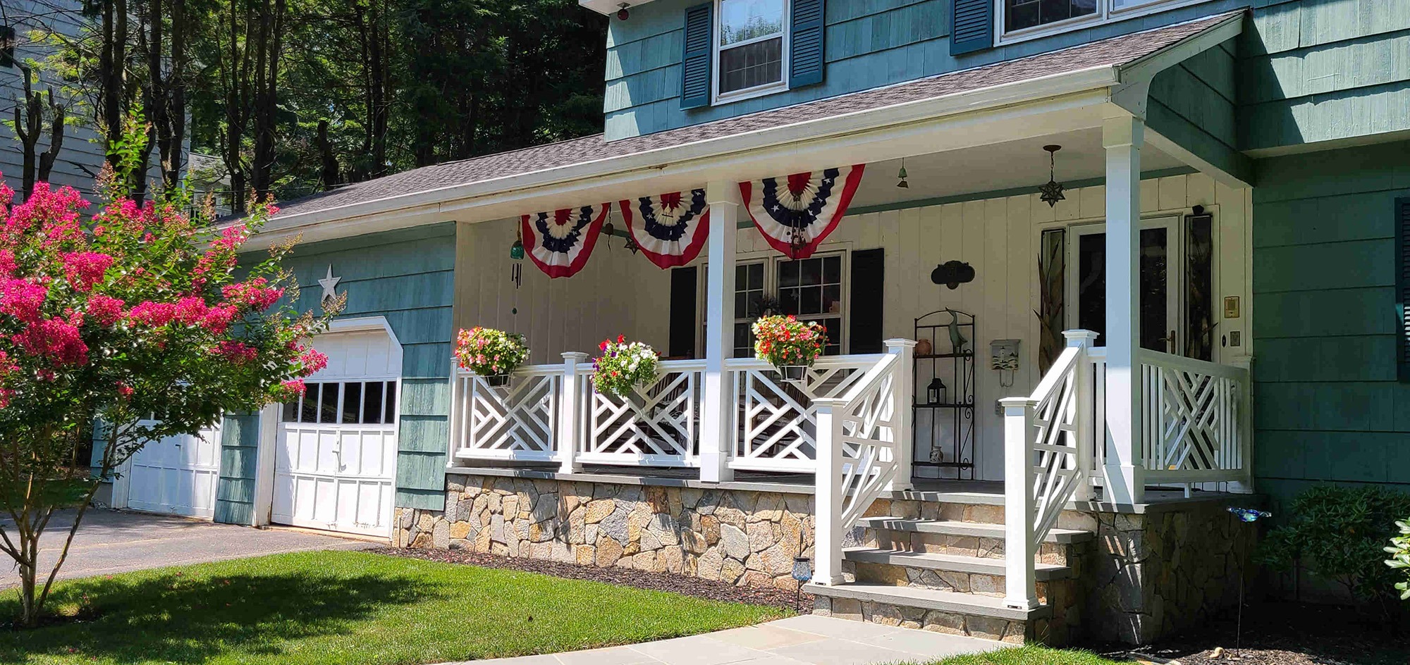 Charming house exterior featuring a cozy porch adorned with red, white, and blue bunting, vibrant flower pots, and a landscaped yard.