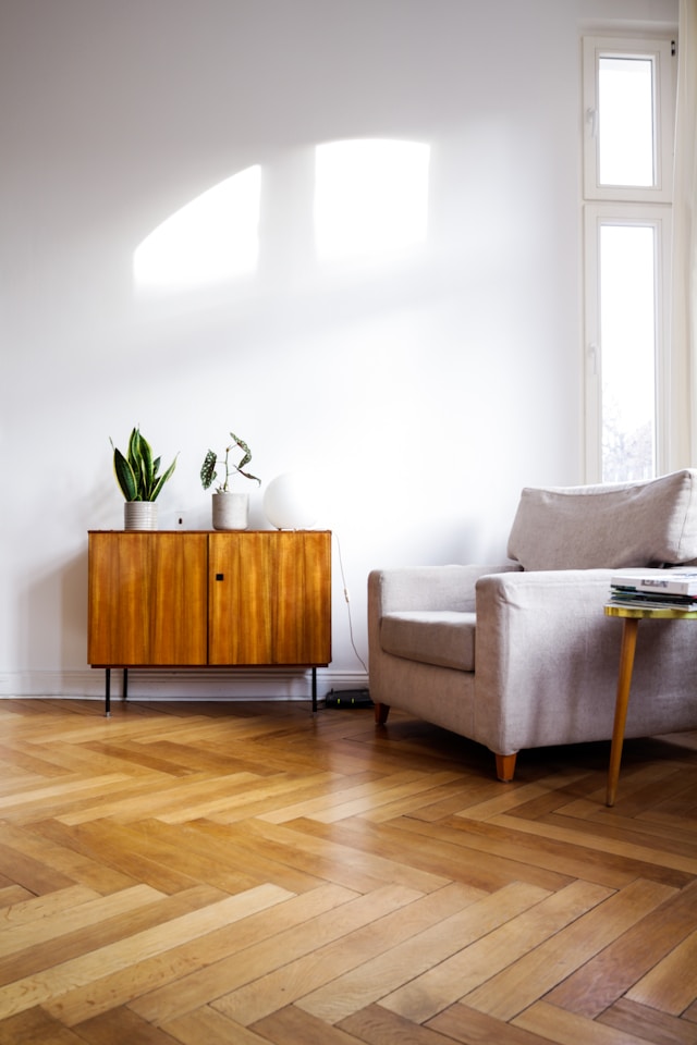 Bright, airy living room featuring a cozy beige armchair, a stylish wooden sideboard with potted plants, and elegant herringbone flooring.