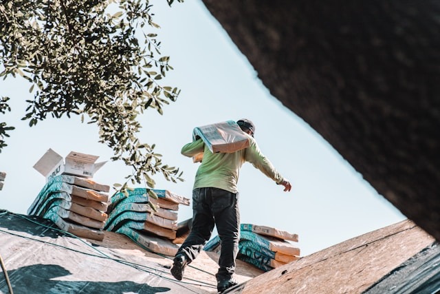 A worker carrying a sack climbs a sloped roof, with stacks of bags visible in the background, highlighting construction activity and labor.