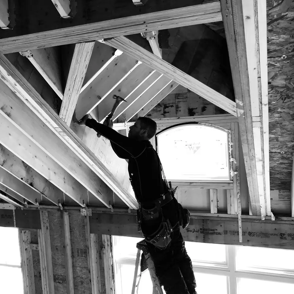 Worker hammering wooden beams while standing on a ladder in a partially constructed building, highlighting ongoing construction efforts.