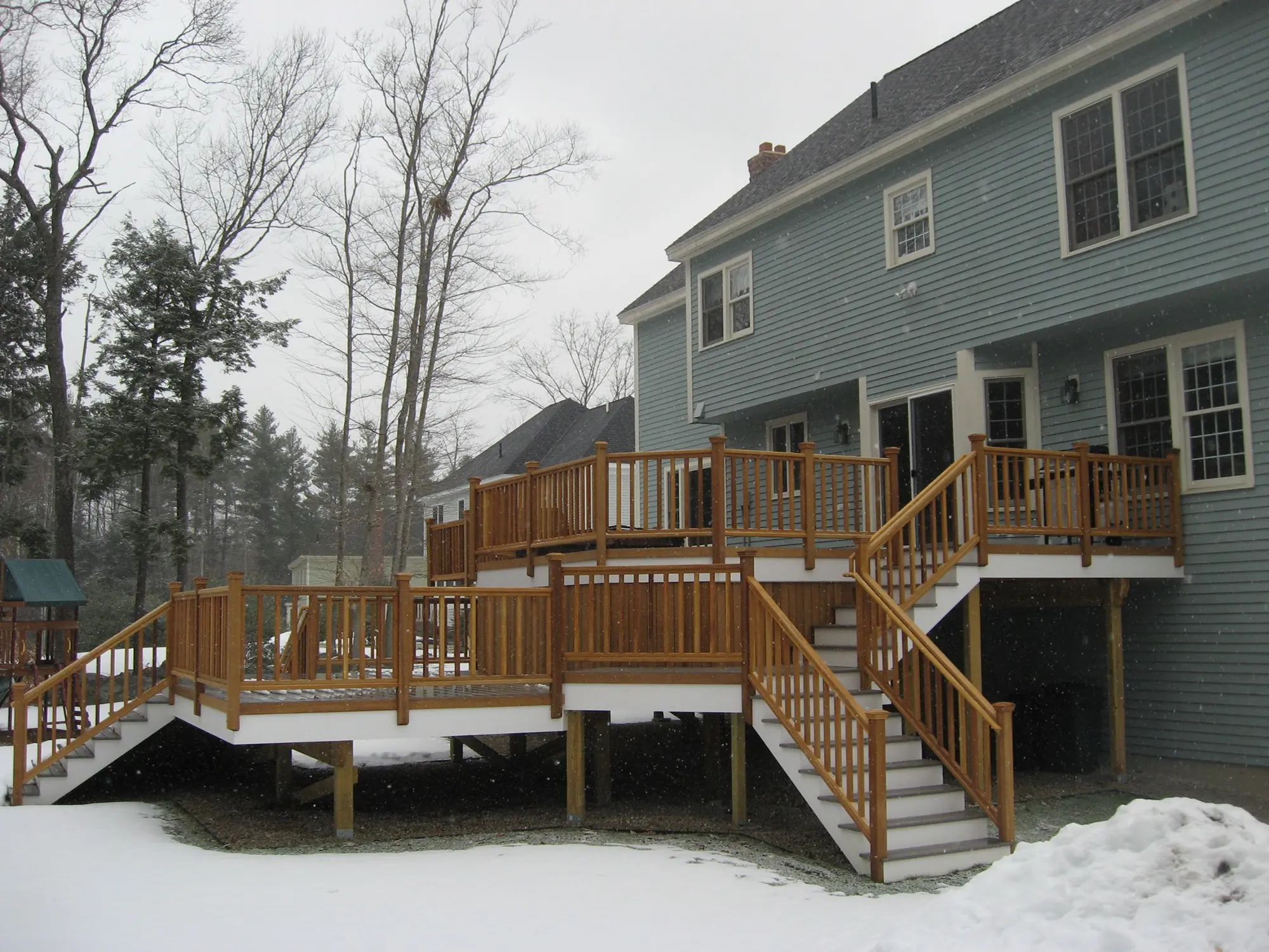 Two-level wooden deck attached to a blue house, surrounded by snow-covered ground and leafless trees, creating a cozy winter outdoor space.