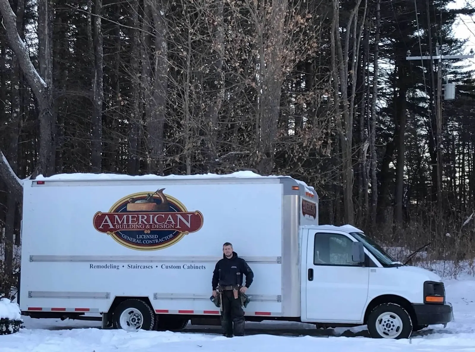 A contractor stands next to an "American Building & Design" truck in a snowy forest setting, showcasing services like remodeling and cabinetry.