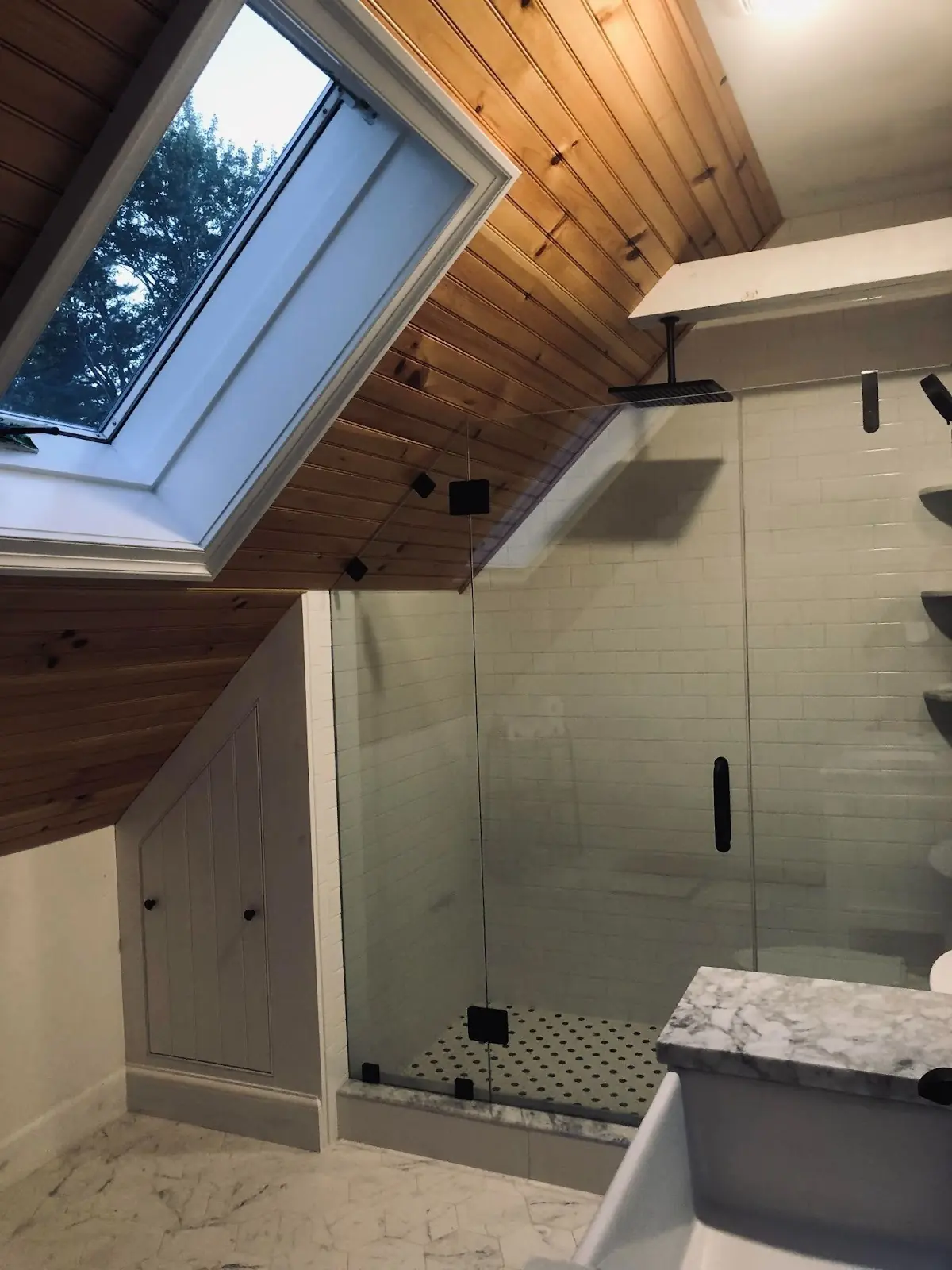 Modern bathroom featuring a skylight, wooden ceiling, and a spacious glass shower with black hardware and white tile accents.