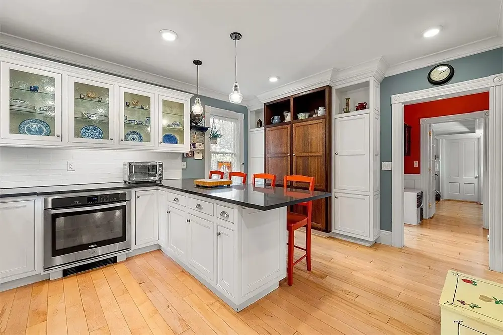 Modern kitchen featuring white cabinetry, a dark countertop, orange stools, and glass-fronted cabinets displaying blue dinnerware. Bright and inviting.