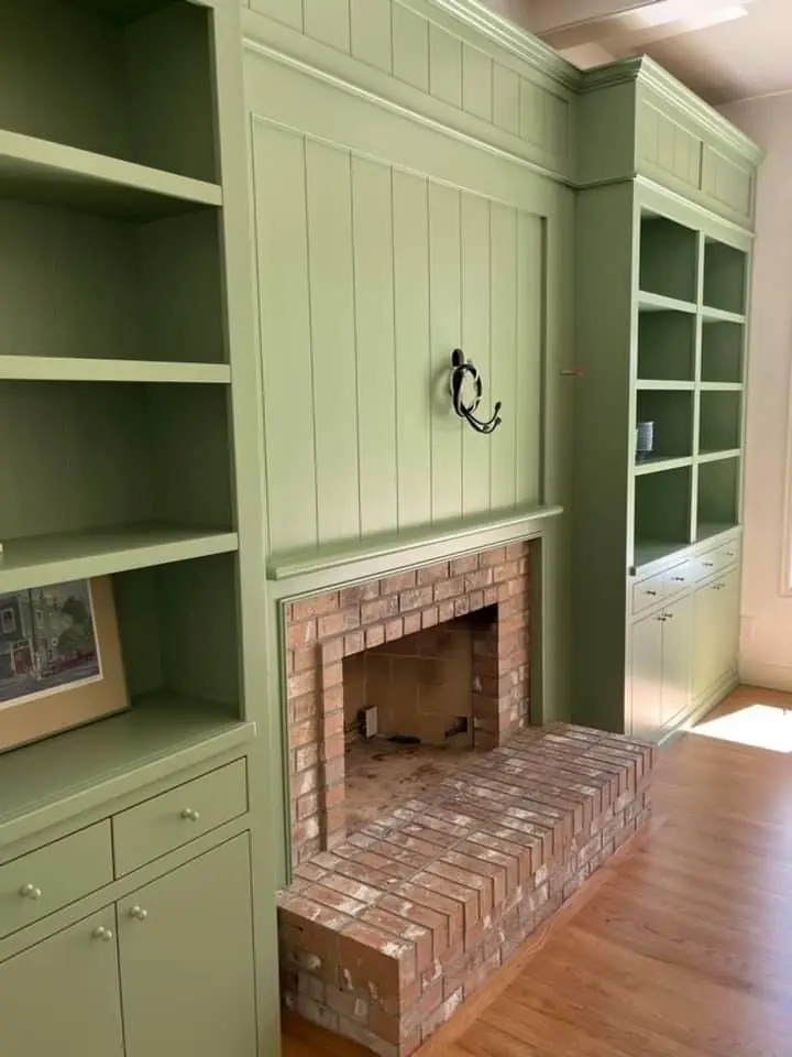 A light green built-in bookshelf frames a small brick fireplace, creating a cozy living space with natural light.