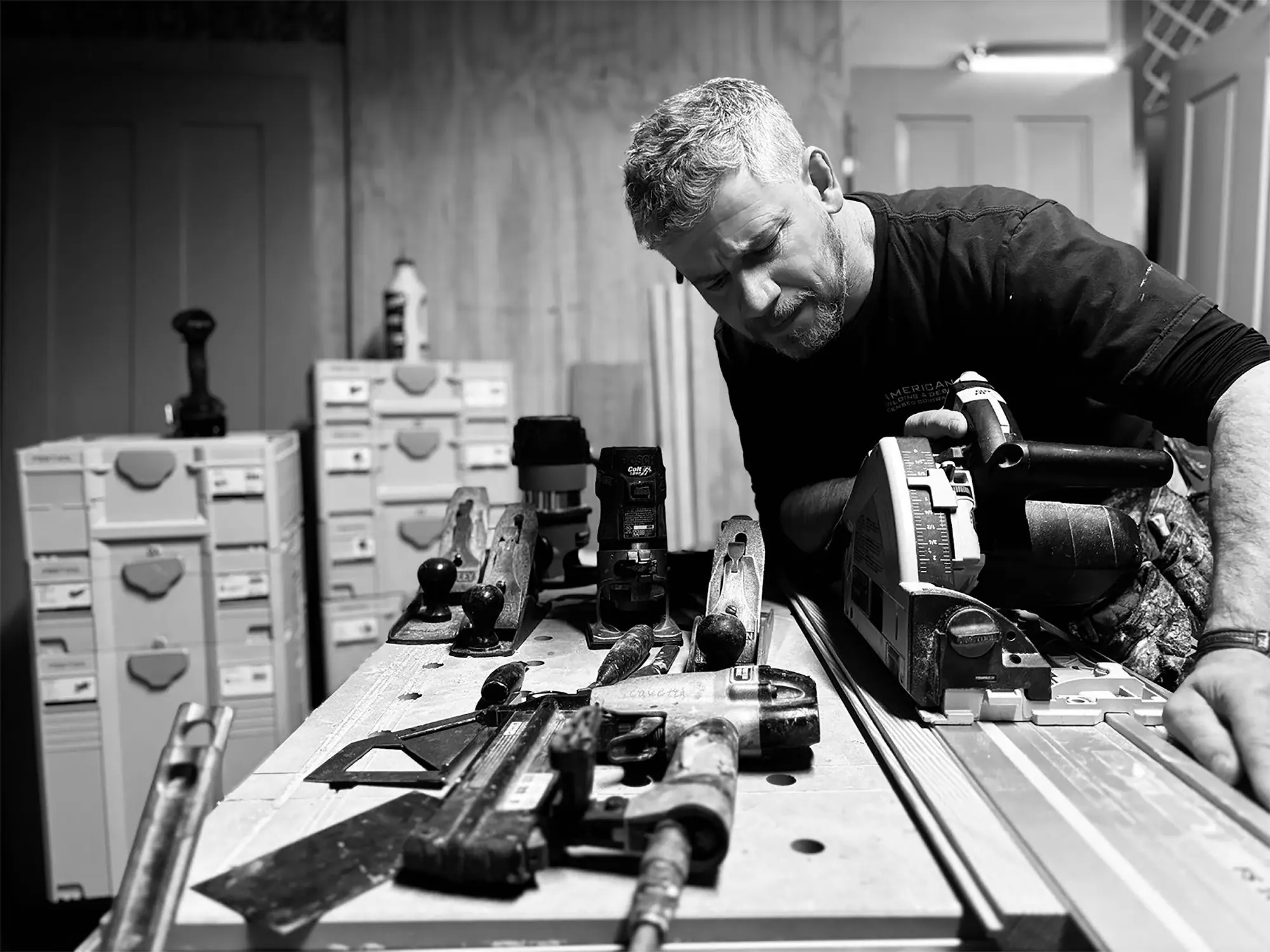A craftsman intensely focuses on using power tools amid an organized workspace, showcasing precision woodworking techniques in a black-and-white setting.