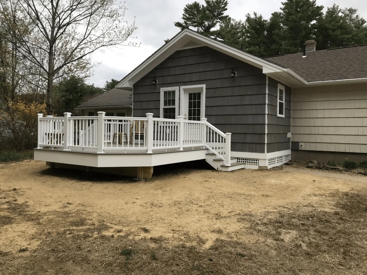 Newly built deck with white railing attached to a gray house, surrounded by bare earth and sparse vegetation, offering outdoor living space.