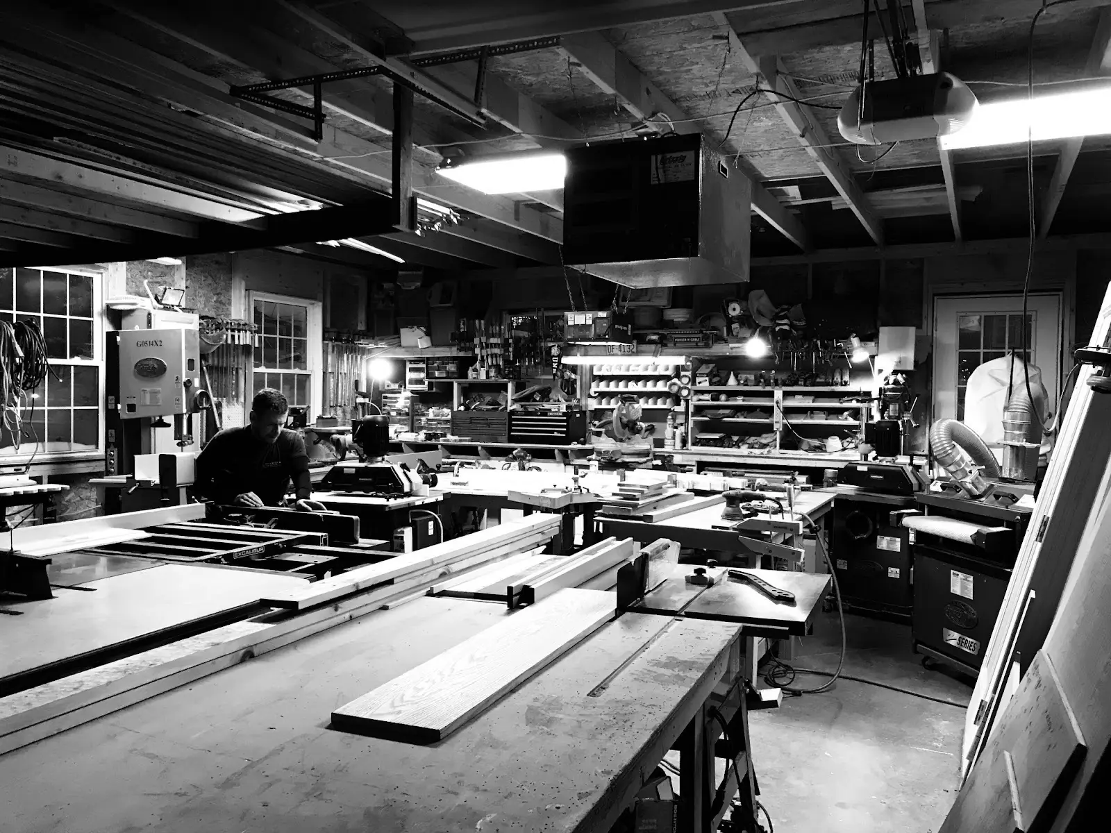 Woodworking shop illuminated by overhead lights, featuring a craftsman focused on a project amidst tools and materials on workbenches.