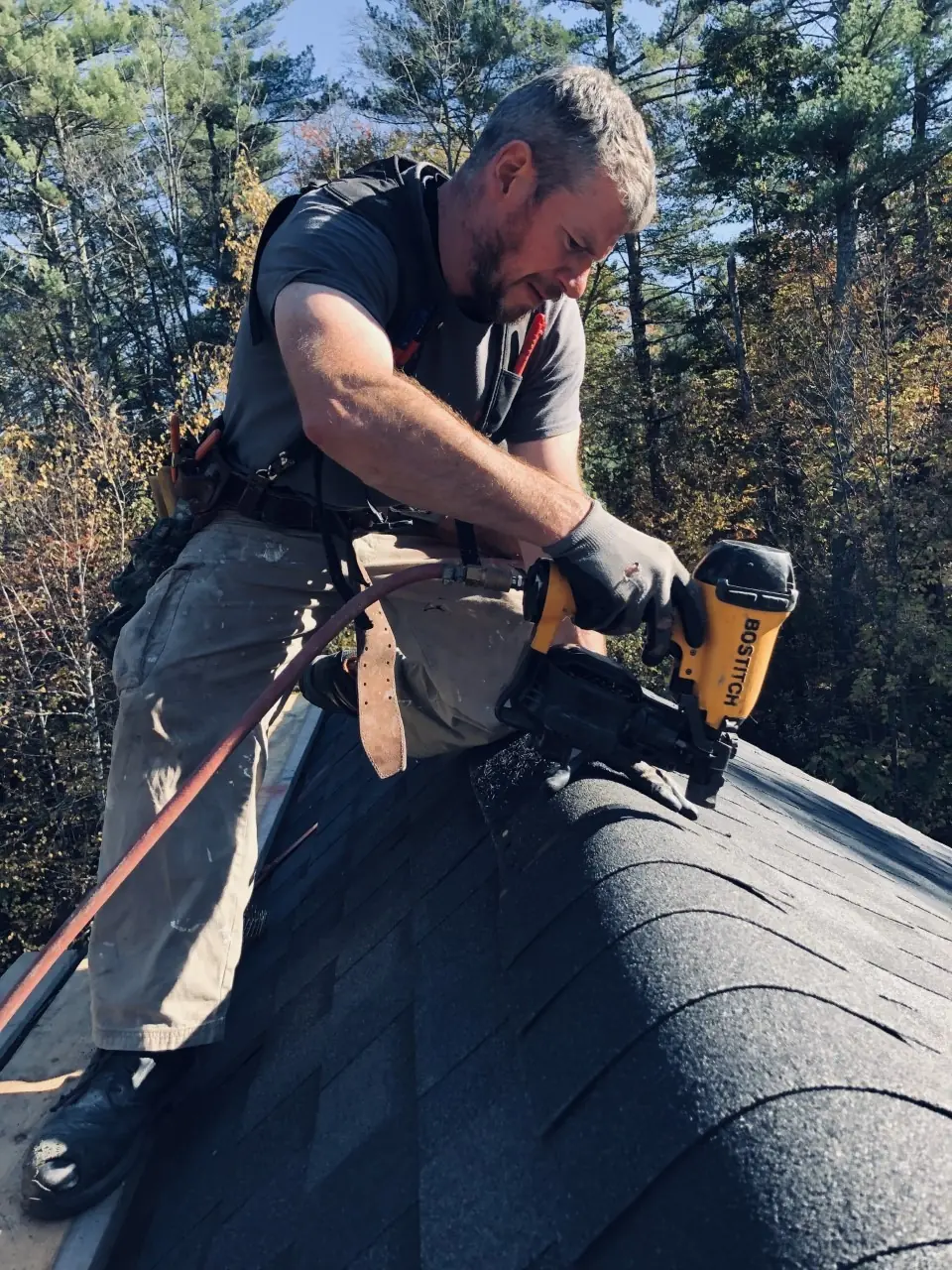Roofer using a nail gun to secure shingles on a sloped roof, surrounded by autumn foliage. The image showcases professional roofing work in progress.
